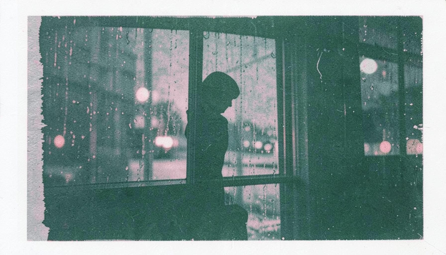 A moody risograph print in muted teal and dusty rose showing a silhouetted figure standing by a rain-streaked window at night, with soft streetlight glow outside.